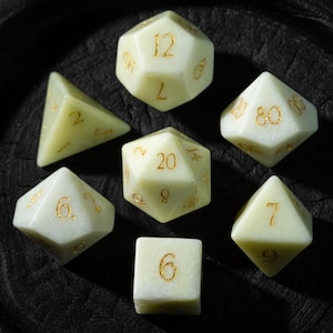 May include: A collection of seven ivory-coloured polyhedral dice with gold numbering. The dice are displayed on a dark, textured surface. The set includes a d4, d6, d8, d10, d12, d20, and a percentile die.
