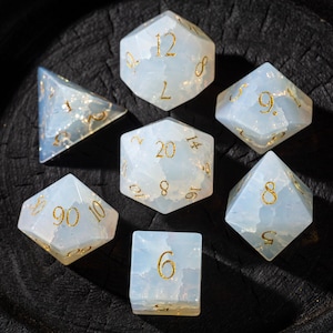 May include: A set of seven polyhedral dice in various shapes, including a d20, d12, d10, d8, d6, and d4. The dice are a translucent, opalescent blue with gold numbering, perfect for tabletop gaming.