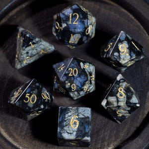 May include: Set of seven polyhedral dice with a speckled black, blue, and gray pattern. The numbers are in gold. The dice are arranged on a dark wooden surface. The dice are for role-playing games.