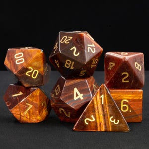 Op de afbeelding: Set van zeven rode tijgeroog-stenen dobbelstenen met gouden cijfers. De dobbelstenen zijn in verschillende vormen, waaronder een d4, d6, d8, d10, d12 en d20.