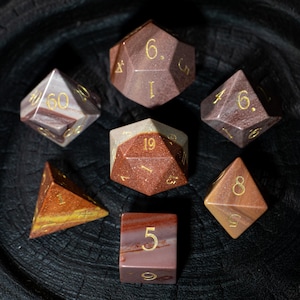 Könnte beinhalten: Eine Sammlung von sieben Edelsteinwürfeln in verschiedenen Formen, darunter ein d4, d6, d8, d10, d12, d20 und ein Würfel. Die Würfel sind hauptsächlich braun und beige mit goldenen Zahlen. Die Würfel sind auf einer dunklen, strukturierten Oberfläche angeordnet.