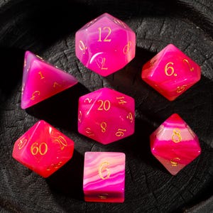 Könnte beinhalten: Ein Satz von sieben polyedrischen Würfeln in Rosa- und Weißtönen mit goldfarbenen Zahlen. Die Würfel bestehen aus einem durchscheinenden Material, und die Zahlen sind gut sichtbar. Die Würfel sind auf einer dunklen Oberfläche angeordnet.