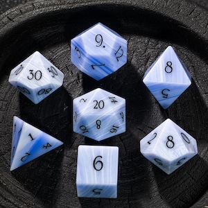 Könnte beinhalten: Set aus sieben blau-weiß marmorierten, polyedrischen Würfeln für Tabletop-Spiele. Die Würfel umfassen einen D4, D6, D8, D10, D12, D20 und einen Prozentwürfel. Jeder Würfel hat schwarze Zahlen.
