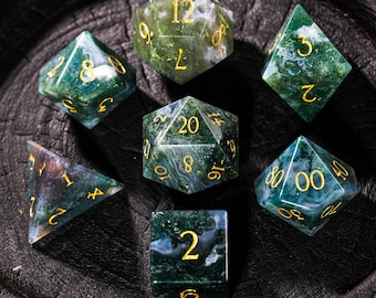 Pode incluir: Conjunto de sete dados poliédricos com um padrão marmoreado verde e branco, com números dourados. Os dados estão dispostos sobre uma superfície escura e texturizada, mostrando várias formas, incluindo um d20, d12, d10, d8, d6 e d4.