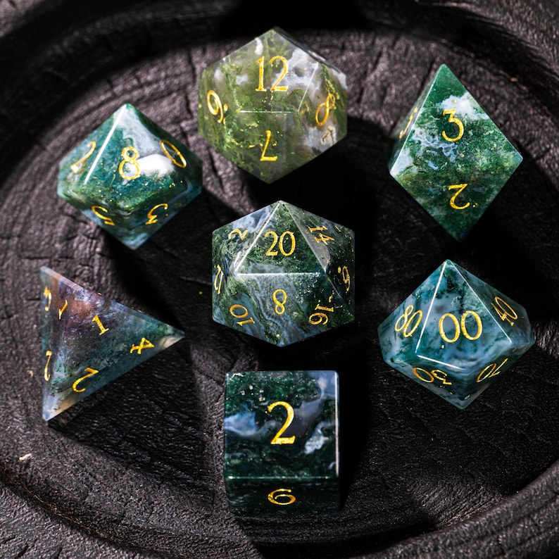 Pu&ograve; includere: Set di sette dadi poliedrici con un motivo marmorizzato verde e bianco, con numeri dorati. I dadi sono disposti su una superficie scura e strutturata, mostrando varie forme tra cui un d20, d12, d10, d8, d6 e d4.