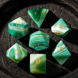 Könnte beinhalten: Ein Set aus sieben grünen und weißen Edelsteinwürfeln mit goldfarbenen Zahlen. Die Würfel haben verschiedene geometrische Formen, darunter ein d4, d6, d8, d10, d12 und d20. Die Würfel liegen auf einer dunklen Holzoberfläche.