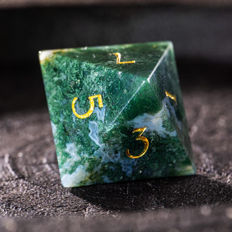 Pu&ograve; includere: Un dado D6 in pietra preziosa verde e bianca con numeri color oro. Il dado ha la forma di una piramide a sei facce, con i numeri 1, 3 e 5 visibili. La pietra ha un aspetto muschioso.