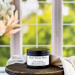 May include: Black candle jar with a white label that says "Your Image Here" and "2 Files Included - High Resolution JPEG - PSD with Smart Object". The candle jar is sitting on a wooden table with a white cloth draped over it. The background is a blurred image of a window with green foliage.
