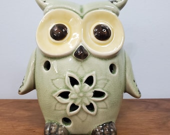 May include: A ceramic owl figurine in shades of green, cream, and brown. The owl has large, round eyes, a flower-shaped cutout on its chest, and several small holes. The figurine is sitting on a wooden surface.