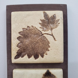 Puede incluir: Adorno de pared con dos baldosas de cer&aacute;mica beige con dise&ntilde;os de hojas en relieve marr&oacute;n, montadas sobre una tabla de madera marr&oacute;n oscuro. Las hojas detalladas contrastan con las baldosas.