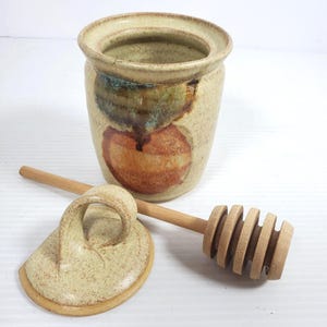 May include: A beige ceramic honey pot with a lid and wooden honey dipper. The pot has a wide opening and is decorated with brown and green abstract shapes. The lid is off to the side, and the dipper rests beside it.