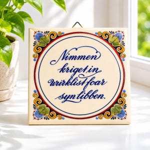 May include: Square ceramic tile with a cream-colored background and a blue and red circular design. The tile features ornate floral corner accents in blue, green, and yellow. The text "Nimmen kriget in wirklisf foar syntibben" is written in blue script.