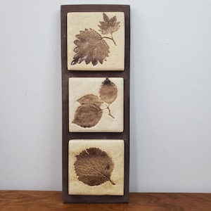 Könnte beinhalten: Ein rechteckiges Wandbild mit drei quadratischen Fliesen, die jeweils einen braunen Blatteindruck zeigen. Die Fliesen sind in einem dunkelbraunen Holzrahmen eingefasst. Die Blätter weisen verschiedene Formen und Texturen auf und wirken natürlich und rustikal.