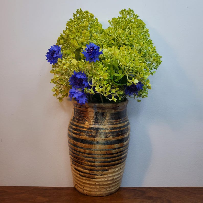 K&ouml;nnte beinhalten: Eine Keramikvase mit braunem und schwarzem Streifendesign h&auml;lt eine lebendige Anordnung aus gr&uuml;nem Laub und blauen Blumen. Die Vase hat eine strukturierte Oberfl&auml;che und eine breite, abgerundete Form. Die Blumen setzen einen Farbtupfer.