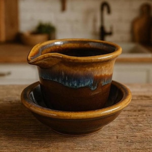 Puede incluir: Un juego de salsera y platillo de cerámica hechos a mano. El bote tiene un pico y un esmalte marrón oscuro con detalles azules y dorados. El platillo es marrón oscuro a juego con ribete dorado. El juego está sobre una superficie de madera.