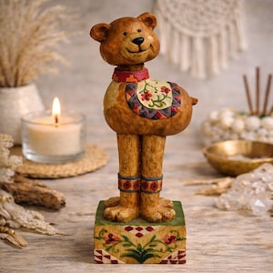 Peut inclure: Figurine d'ours brun fantaisiste avec un visage souriant, portant un collier et une selle décoratifs. L'ours est posé sur une base verte avec des motifs floraux. La figurine présente des détails et est probablement en résine.
