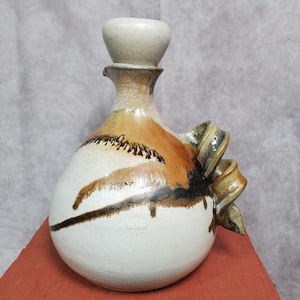 May include: A handmade ceramic decanter with a bulbous body and a small spout. The decanter features a cream-colored base with brown and black brushstrokes and a decorative handle. The stopper is a light beige color.