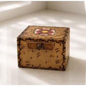 May include: A square wooden box with a light brown finish, featuring a hand-painted pink and yellow flower on the lid. The box has a dark brown scalloped border design along the edges and a small, centred wooden knob on the front panel.
