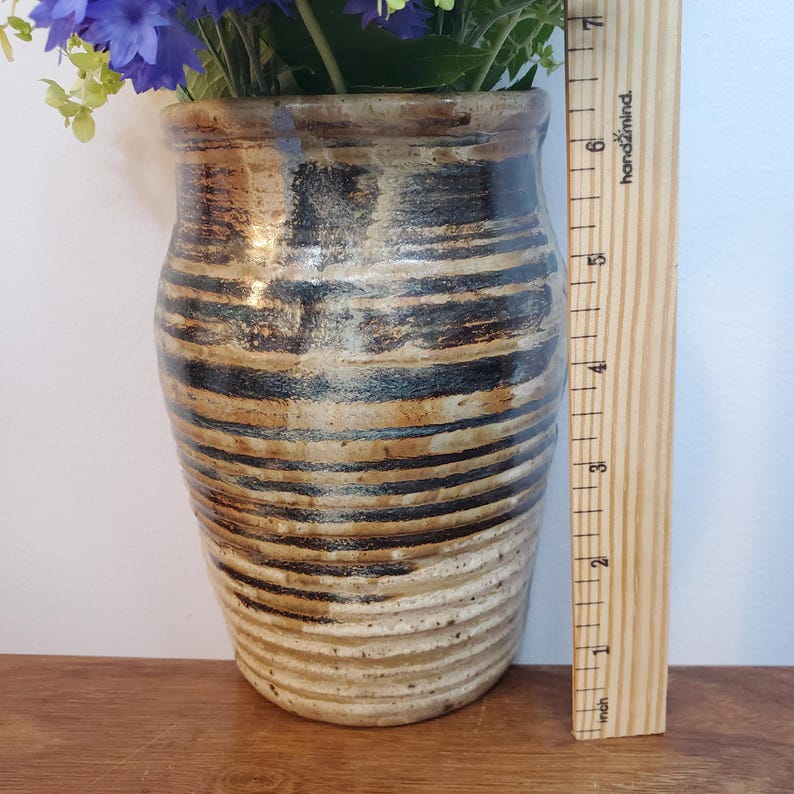 K&ouml;nnte beinhalten: Handgefertigte Keramikvase mit Streifen in Braun-, Schwarz- und Beiget&ouml;nen, gef&uuml;llt mit blauen und gr&uuml;nen Blumen. Die Vase ist etwa 18 cm hoch, wie das Holzlineal daneben zeigt. Rustikales, handgemachtes Aussehen.