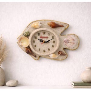 Könnte beinhalten: Eine fischförmige Wanduhr mit sandigem Hintergrund und Muscheln. Das Zifferblatt ist beige mit schwarzen Zahlen und Zeigern. Die Uhr ist von verschiedenen Muscheln in Beige-, Braun- und Weißtönen umgeben.