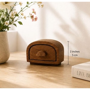 May include: A small, arched wooden storage box with a single drawer and a semi-circular handle. The box features a natural brown wood grain finish and stands 5 cm tall, as indicated by the measurement text printed next to the object.