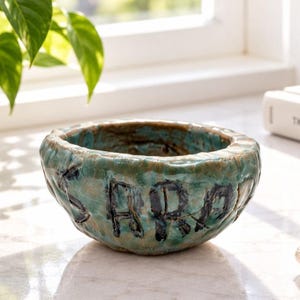 Peut inclure: Un petit bol en céramique de forme ronde, réalisé à la main, présentant une surface texturée et irrégulière. L'émail est un mélange tacheté de tons turquoise, bleu sarcelle et brun terreux. Les lettres SARA sont gravées sur le côté extérieur.
