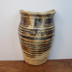 K&ouml;nnte beinhalten: Eine handgefertigte Keramikvase mit einem einzigartigen Streifendesign. Die Vase hat horizontale B&auml;nder in Braun-, Schwarz- und Beiget&ouml;nen. Der obere Rand hat eine geschwungene Form. Die Vase ist ein dekorativer Artikel f&uuml;r die Inneneinrichtung.