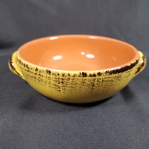 Puede incluir: Un cuenco de cerámica con exterior amarillo y diseño moteado marrón. El interior es naranja. El cuenco tiene dos asas pequeñas y es adecuado para servir sopas, guisos u otros platos.