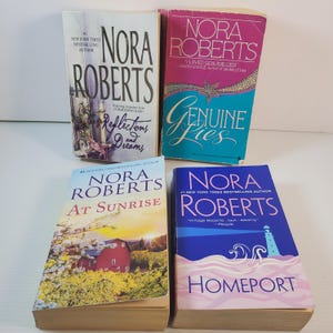 May include: Four paperback books by Nora Roberts are shown. The titles are "Reflections and Dreams", "Genuine Lies", "At Sunrise", and "Homeport". The covers have diverse designs and colours, with the author's name clearly visible.