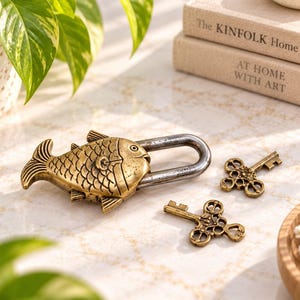 May include: A brass-colored metal padlock shaped like a fish with a dark, weathered steel shackle. Two ornate, matching brass-colored skeleton keys with intricate circular bow designs are positioned next to the fish-shaped padlock on a light surface.