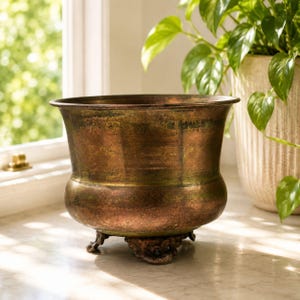 Op de afbeelding: Een verweerde bronzen plantenbak met een brede, uitlopende rand en een afgeronde body. De plantenbak rust op drie sierlijke poten. Een groene plant is zichtbaar op de achtergrond, wat suggereert dat de plantenbak decoratief gebruikt wordt.