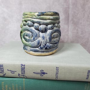 May include: A handmade ceramic mug with a unique, textured design. The mug features a blue and green color scheme with raised, organic shapes. The base is a light tan color. The mug is sitting on top of two books.