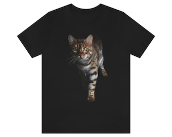 May include: A black t-shirt featuring a brown and black tabby cat walking towards the camera. The cat has green eyes and is in a dark setting.