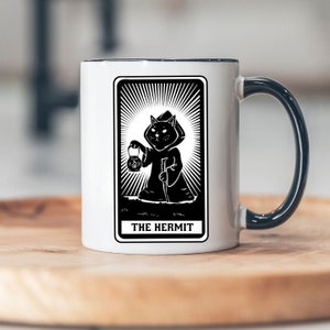 May include: White ceramic mug with a black handle. The mug features a black and white illustration of a cat dressed as The Hermit from a tarot deck. The text "THE HERMIT" is printed below the image.