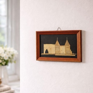 Peut inclure: Une œuvre d'art encadrée représentant un château. L'œuvre présente une structure de château brun clair sur un fond noir, encadrée dans un cadre en bois brun foncé. Le château a deux hautes tours et une entrée en arc.