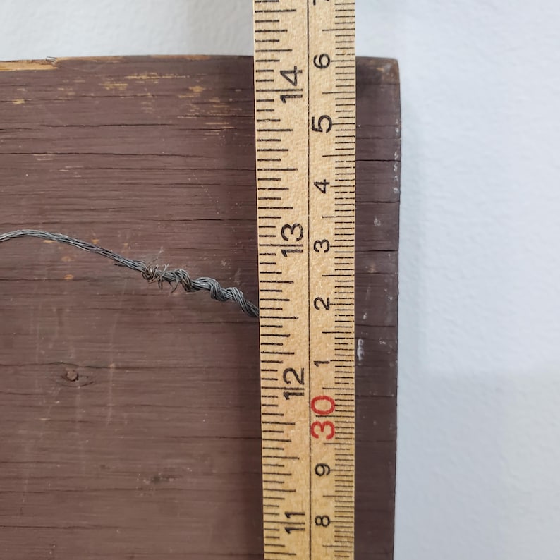 Puede incluir: Una regla de madera con marcas en pulgadas se coloca junto a una tabla de madera marr&oacute;n. La regla muestra medidas de hasta 35,5 cm. Un alambre est&aacute; sujeto a la tabla de madera.