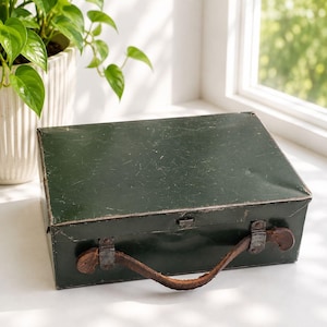 May include: A rectangular dark green metal storage box with a hinged lid and a worn brown leather handle attached by metal brackets. The box features a smooth, slightly scratched surface and a simple metal latch mechanism on the front side.