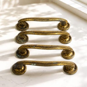 Puede incluir: Cuatro tiradores de cajón antiguos de latón. Cada tirador presenta un asa curvada con una textura de cuerda y placas de extremo circulares decorativas. El latón tiene una pátina cálida y envejecida, lo que sugiere herrajes antiguos. Ideal para la restauración de muebles.