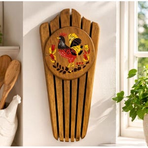 Könnte beinhalten: Küchenutensilienhalter aus Holz mit Hahnmotiv. Der Halter besteht aus vertikalen Holzlatten mit einem kreisförmigen Dekorationselement mit einer Hahnillustration in Rot, Gelb und Schwarz. Er ist an einer weißen Wand befestigt.