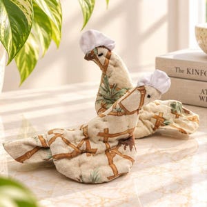 May include: Two quilted fabric oven mitts shaped like chickens, featuring a beige background with a brown bamboo-style fence pattern and green foliage accents. Each chicken-shaped mitt is topped with a small, white, gathered fabric chef hat.