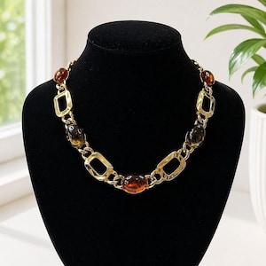 May include: A gold-coloured necklace featuring amber-coloured oval and rectangular links. The necklace is displayed on a black mannequin. The necklace has a chain-like design with a mix of oval and rectangular amber-coloured stones.
