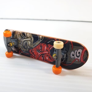Puede incluir: Un monopatín en miniatura con una tabla negra con diseños gráficos rojos y dorados de caras. Las ruedas son naranjas y los ejes grises. Diseñado para fingerboarding.