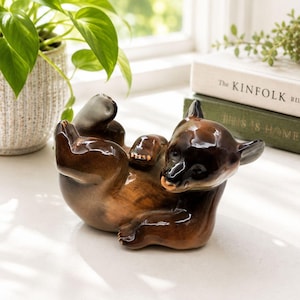 Peut inclure: Une figurine en céramique brillante représentant un ourson brun allongé sur le dos. L'ours a une tête brun foncé et un corps brun plus clair avec des pattes et des griffes visibles. La sculpture présente une finition lisse et réfléchissante.