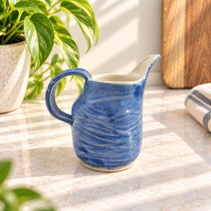 Peut inclure: Un pichet à crème en céramique fait main présentant une glaçure bleue vibrante avec une surface texturée et ondulée. Le pichet possède une anse incurvée sur un côté et un bec verseur pointu, avec une finition intérieure lisse blanc cassé.