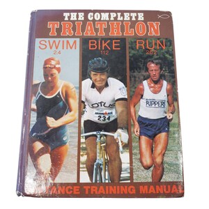 May include: A vintage book titled "THE COMPLETE TRIATHLON" with images of a swimmer, cyclist, and runner. The cover features the distances for each sport: 2.4 miles swim, 112 miles bike, and 26.2 miles run. The book is a performance training manual.