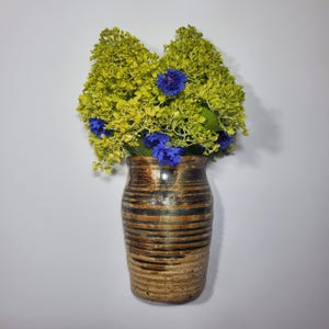 K&ouml;nnte beinhalten: Eine Keramikvase mit braunem und schwarzem Streifenmuster h&auml;lt einen Strau&szlig; gelb-gr&uuml;ner und leuchtend blauer Blumen. Die Vase hat ein rustikales, handwerkliches Aussehen, ideal f&uuml;r die Inneneinrichtung.