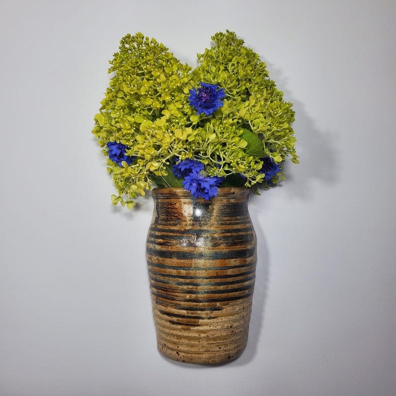 K&ouml;nnte beinhalten: Eine Keramikvase mit braunem und schwarzem Streifenmuster h&auml;lt einen Strau&szlig; gelb-gr&uuml;ner und leuchtend blauer Blumen. Die Vase hat ein rustikales, handwerkliches Aussehen, ideal f&uuml;r die Inneneinrichtung.