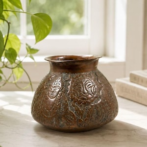 Op de afbeelding: Een ronde koperen vaas met een brede opening en een taps toelopende hals. Het buitenoppervlak is versierd met reliëfpatronen van gestileerde menselijke gezichten en bloemmotieven, afgewerkt met een donkere, verweerde patina.