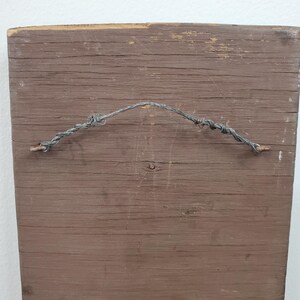 Puede incluir: Parte trasera de un letrero rectangular de madera con acabado marr&oacute;n. Se adjunta un alambre retorcido para colgarlo. La veta de la madera es visible y el letrero muestra signos de desgaste y edad.