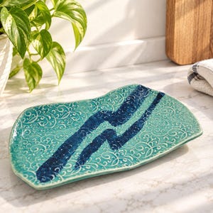 May include: A rectangular ceramic tray with an irregular, wavy edge. The surface features a light turquoise glaze with a white swirling filigree pattern and two prominent, dark navy blue diagonal stripes across the centre of the piece.